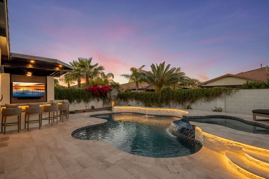 Freeform pool with lit step entry beside the covered bar with TV